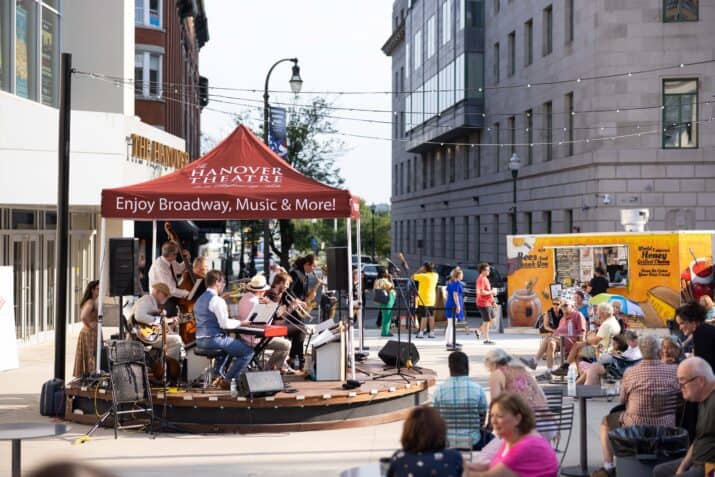 On the left of the photograph, a live band plays on top of the Bank of America stage in front of the Hanover Theatre. In the right foreground, an audience sits and enjoys the music as a Bees And Thank You food truck services others in the background at the Carroll Plaza Ribbon Cutting, 2023. Photo by Unity Mike Photography.