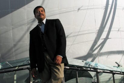 Neil deGrasse Tyson strikes a slanted standing pose in front of a science museum exhibit.