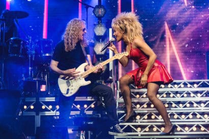 Karis Anderson (right) as Tina Turner in TINA - The Tina Turner Musical. She's wearing a red leather dress and she's singing into a microphone as she leans towards a guitarist. Photo credit to Matt Crockett.