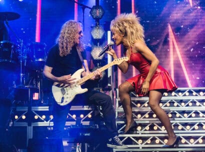 Karis Anderson (right) as Tina Turner in TINA - The Tina Turner Musical. She's wearing a red leather dress and she's singing into a microphone as she leans towards a guitarist. Photo credit to Matt Crockett.