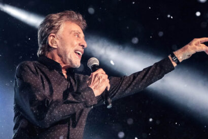 Frankie Valli (of Frankie Valli & The Four Seasons: The Last Encores) looks offstage and off-camera as a white spotlight shines diagonally behind him. He holds his mic close to his mouth in one hand while the other points off in the distance.