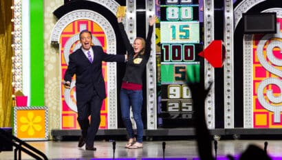 The Price is Right Live host (black suit, purple tie) celebrates with a contestant (black cardigan, blue jeans) with her arms up in front of the game show set and a cheering audience.