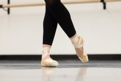 A ballet dancer's legs in black leggings and pointe shoes, captured mid-dance in a studio with a mirrored wall and barres for balance.