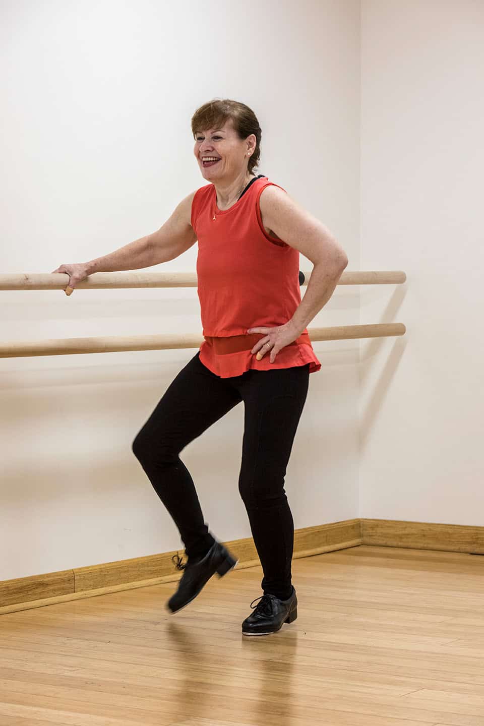 A woman in a red sleeveless top and black pants is tap dancing in a dance studio. She is smiling and balancing with one hand on a ballet barre, with one leg slightly lifted. The room has light-colored walls and wooden flooring.
