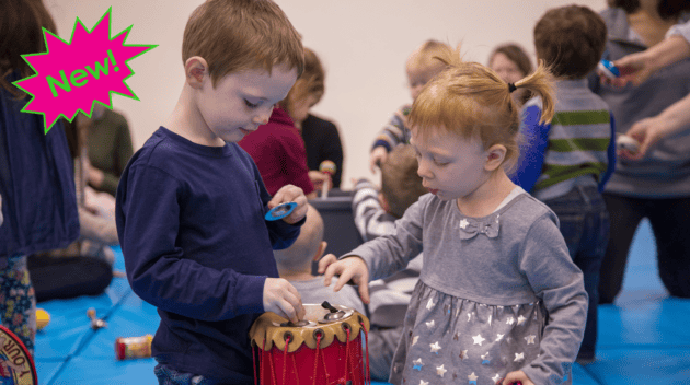 Candid photo of two young students in the Building Reading Comprehension workshop. A boy in navy blue (left) places tiny cymbals on a red drum while a girl in grey (right) reaches for the instrument. In the upper left corner is a neon pink and green exclamation bubble with text inside: "New!"