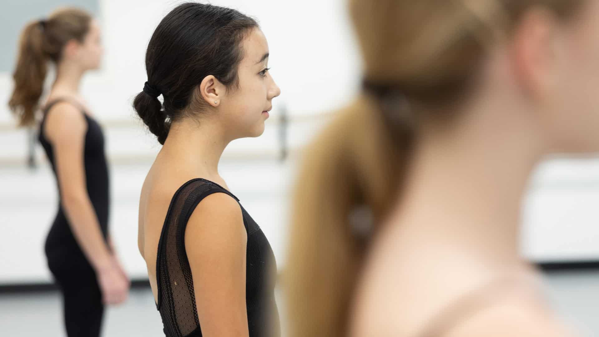 Three young ballet dancers stand in a row, facing to the right. The dancer in the middle, with dark hair pulled back, is in focus, while the other two, one with brunette hair and the other with blonde hair, are blurred. All wear black leotards.