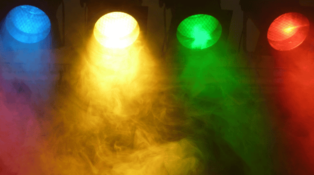 From left to right: blue, yellow, green, and red stage lights shine down on foggy air.