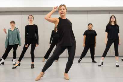 An instructor leads a group of students in a tap dance class. The students wear various casual outfits and are positioned in a dance studio with a light background.