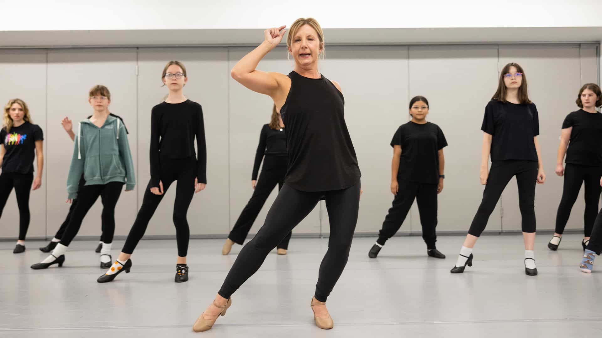 An instructor leads a group of students in a tap dance class. The students wear various casual outfits and are positioned in a dance studio with a light background.