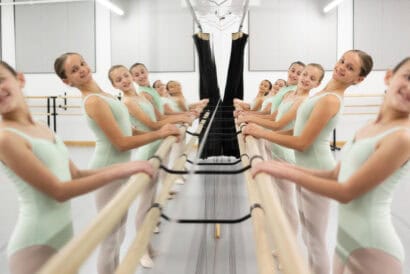 A group of young ballet dancers wearing light blue leotards stands at a barre, practicing their form. They are arranged in a mirrored studio, creating a symmetrical reflection of their aligned positions and graceful poses.