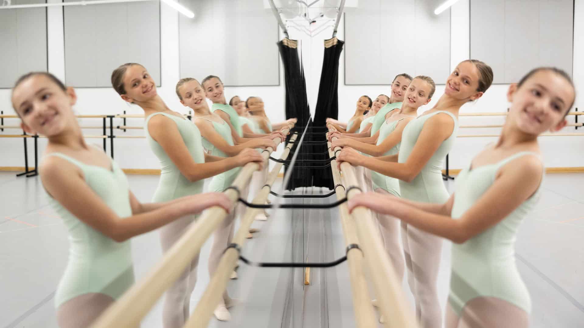 A group of young ballet dancers wearing light blue leotards stands at a barre, practicing their form. They are arranged in a mirrored studio, creating a symmetrical reflection of their aligned positions and graceful poses.