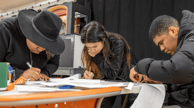 Promotional photo for The Math Behind Scenic Design workshop. Three students all wearing black are looking down, heavily focused on their projects on the table in front of them.