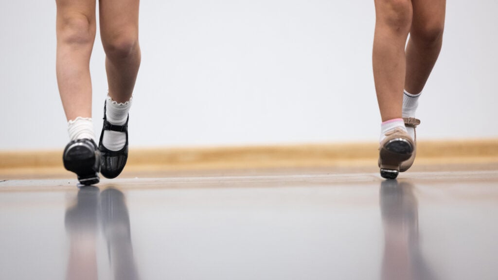 Two children are seen from the knees down, wearing tap shoes and stepping on a smooth surface. One child wears black shoes with frilled socks, and the other wears beige shoes with plain white socks. The background is simple with a light-toned floor and wall.