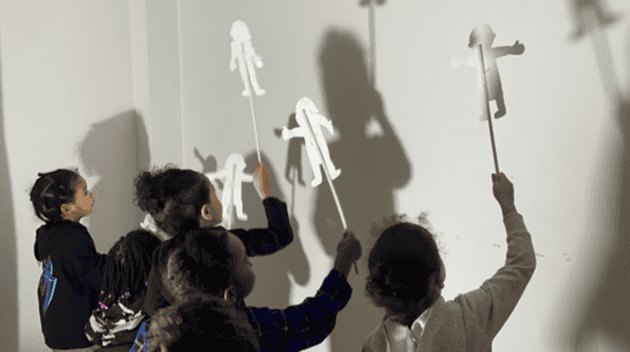 Promotional photo for Lights Up With Shadow Puppets workshop. Photo is lit with a white light and taken from a quarter perspective behind young students holding up sticks with paper figures attached.