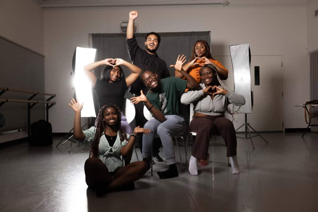 A staged photo of six WYSH 2024 participants smiling with various poses in front of a black fabric backdrop and two large softbox lights in a Conservatory rehearsal room.