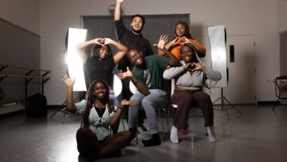 A staged photo of six WYSH 2024 participants smiling with various poses in front of a black fabric backdrop and two large softbox lights in a Conservatory rehearsal room.