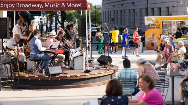 On the left of the photograph, a live band plays on top of the Bank of America stage in front of the Hanover Theatre. In the right foreground, an audience sits and enjoys the music as a Bees And Thank You food truck services others in the background at the Carroll Plaza Ribbon Cutting, 2023. Photo by Unity Mike Photography.