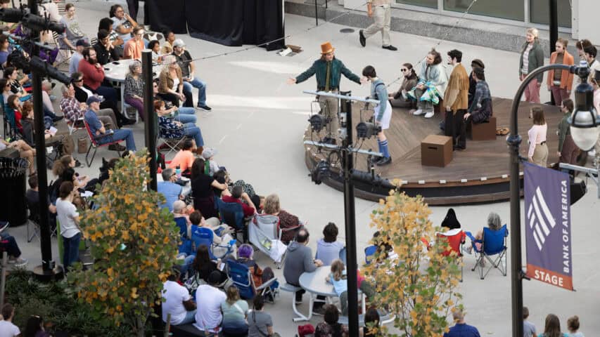 A distant overhead photograph of the Youth Acting Company presenting James and the Giant Peach, 2023-2024. In the foreground are trees with green and yellow leaves, slightly obscuring the audience sitting in a semicircle in front of the Bank of America Stage where the actors are performing. Photo by Unity Mike Photography.