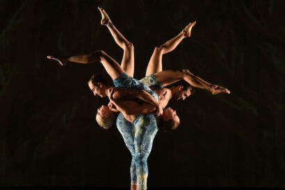 MOMIX: Alice. Two dancers carry two other dancers upside down on their backs in a single file line so that their legs in the air form the shape of a flower.