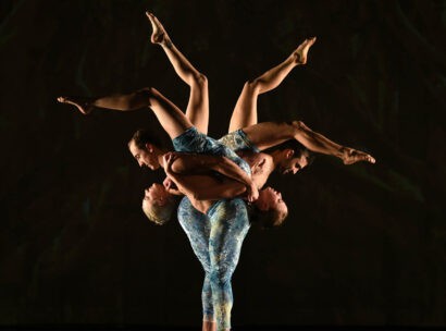 MOMIX: Alice. Two dancers carry two other dancers upside down on their backs in a single file line so that their legs in the air form the shape of a flower.