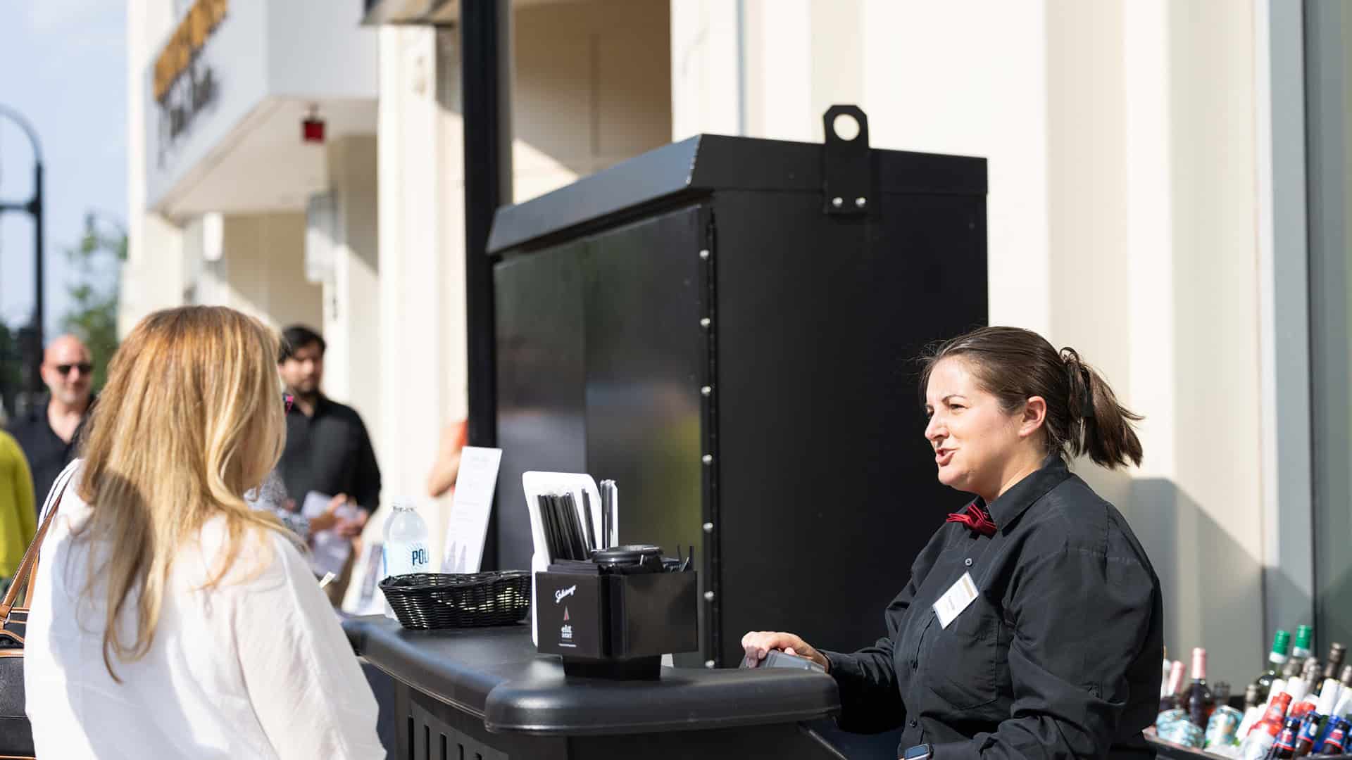A bartender in a red bow tie and black dress shirt at their outdoor station (right) speaking to a customer in white (left) at the Curtain Call Beer and Wine Garden.