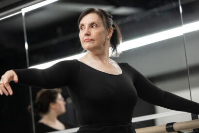 A dancer wearing a black long sleeve leotard in Adult Ballet Beginner II (Ages 18+) looks off-camera as she stretches one arm out and grasps the barre with the other.