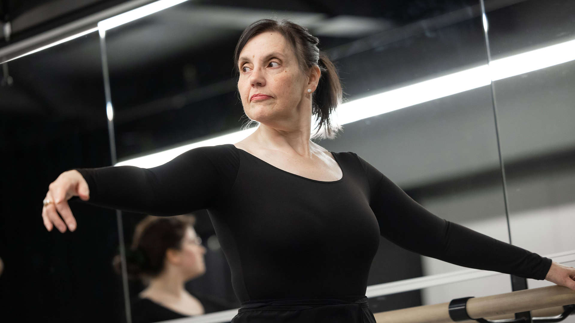 A dancer wearing a black long sleeve leotard in Adult Ballet Beginner II (Ages 18+) looks off-camera as she stretches one arm out and grasps the barre with the other.