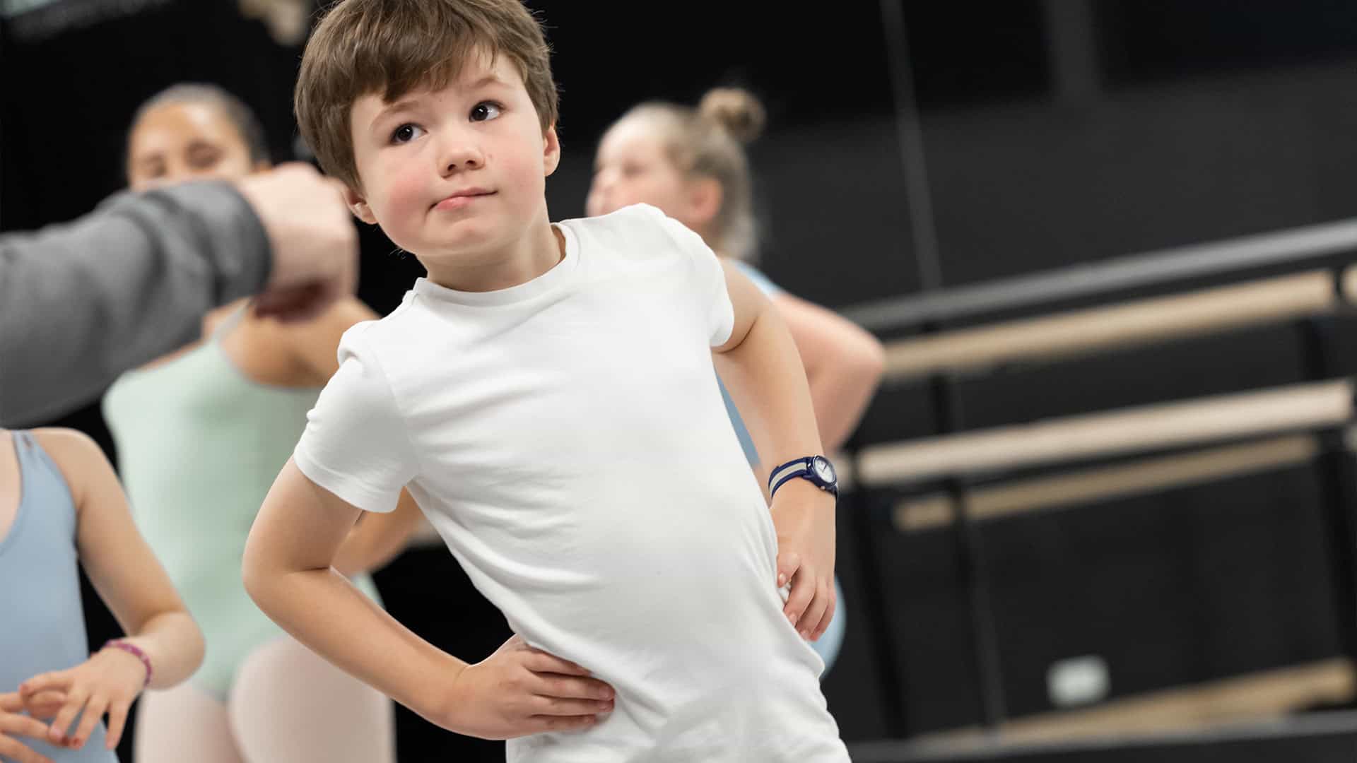 Off camera, an instructor in a grey long sleeve points to a singular male student in Boys Ballet (Ages 8-18). In the background and less in focus are three female ballet students.