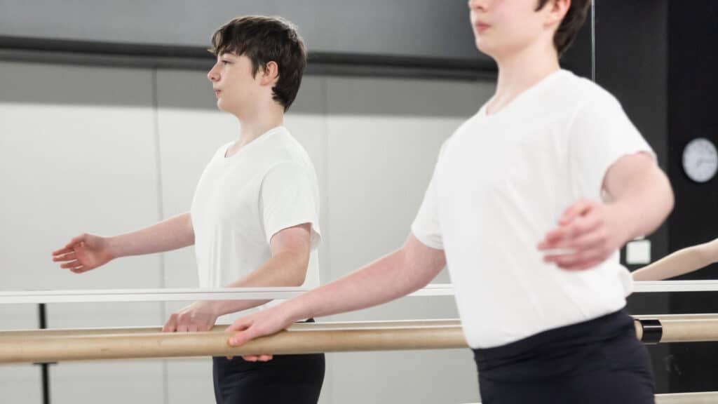 A male student stretches next to his reflection at the dance studio barre in Boys Ballet (Ages 8-18)