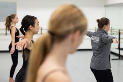 A Dance Performance (Ages 10-14) instructor in a grey branded jacket teaches a dance move in front of three students with the one in the foreground out of focus as she's closest to the camera.