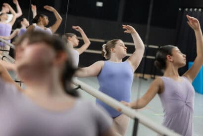 Nine ballet students in Primary Ballet II (Ages 10+) stretch at the dance studio's barre, one arm up each. One student is out of focus in the left foreground.