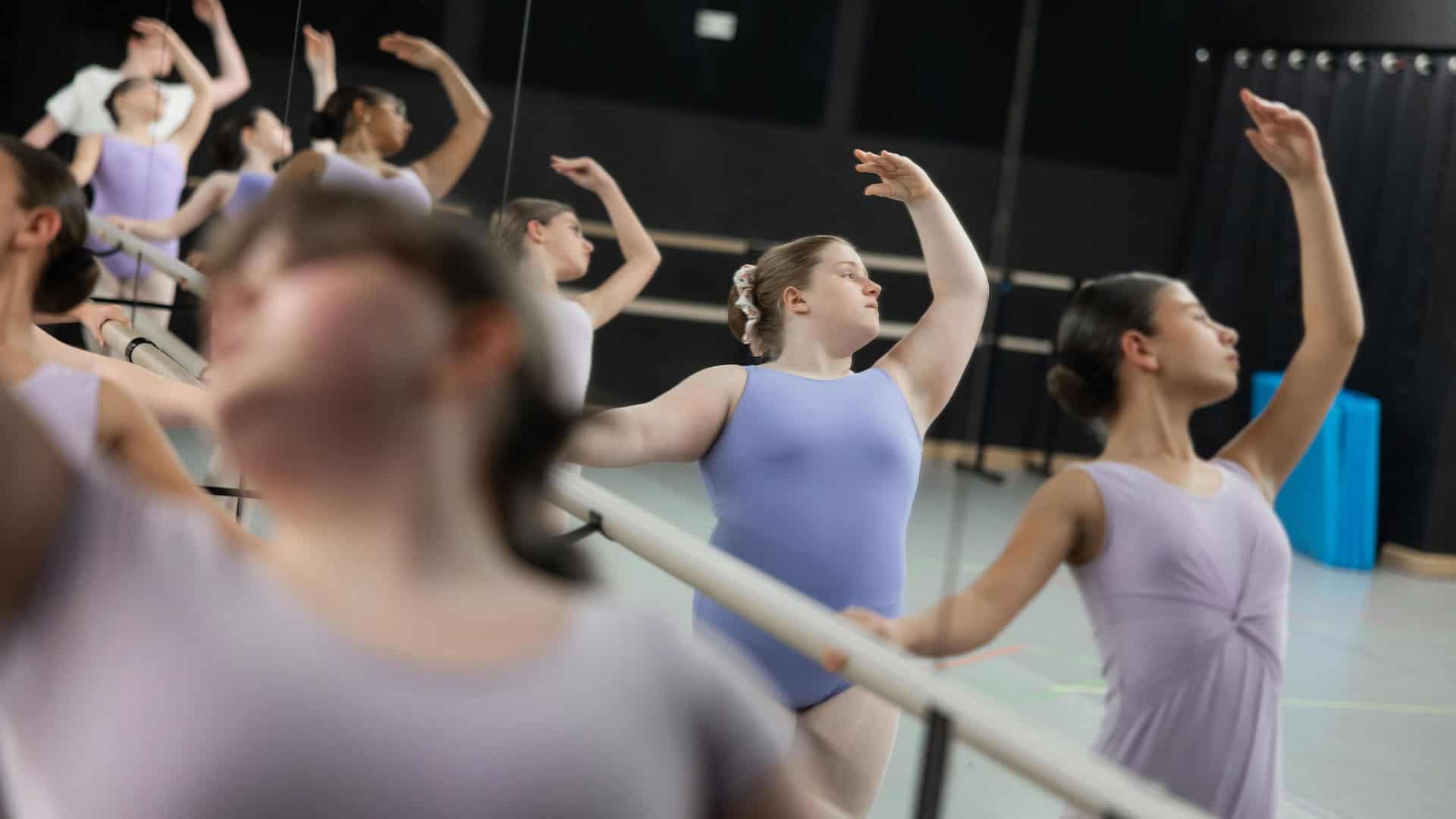 Nine ballet students in Primary Ballet II (Ages 10+) stretch at the dance studio's barre, one arm up each. One student is out of focus in the left foreground.