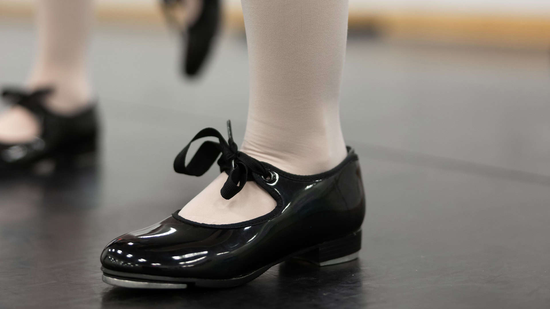 A close up shot of a tap dancer's foot in Primary Tap I (Ages 7-9). She wears pale tights and shiny black tap shoes, the other foot out of focus to the back left.