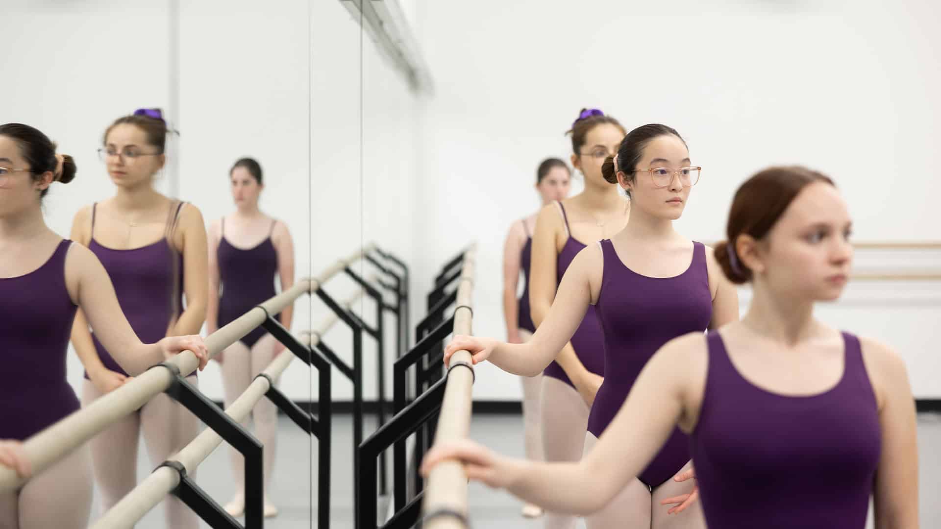 In Teen Ballet Beginner (Ages 12-18), a front facing shot of four ballerinas in purple leotards lined up single file. All students are facing off-camera and two are grasping the dance studio's barre.