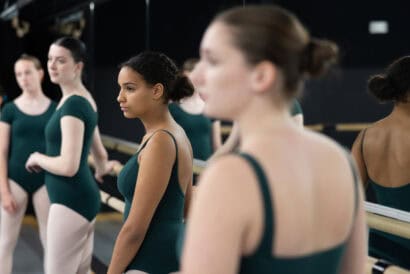 Five ballerinas in the Teen Ballet Beginner (Ages 12-18) class wear green leotards as they look off-camera, lined up against the dance studio's barre. The ballerina closest to the camera has her back turned and is out of focus.