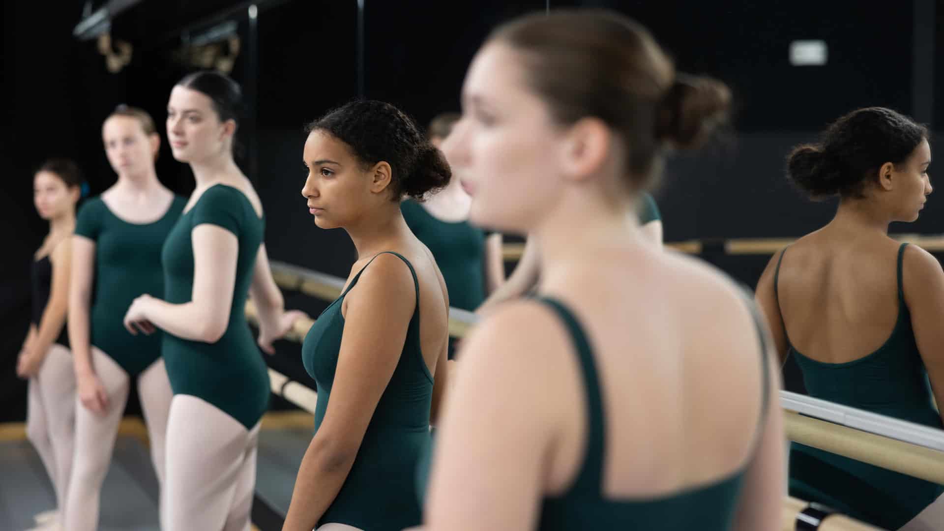 Five ballerinas in the Teen Ballet Beginner (Ages 12-18) class wear green leotards as they look off-camera, lined up against the dance studio's barre. The ballerina closest to the camera has her back turned and is out of focus.