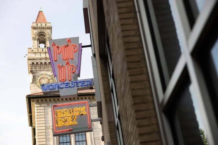 Neon signs for 'PopUp Worcester' and 'BrickBox Worcester' hang on the side of a brick building, reflecting in nearby windows. In the background, Worcester City Hall’s clock tower stands against a clear sky.