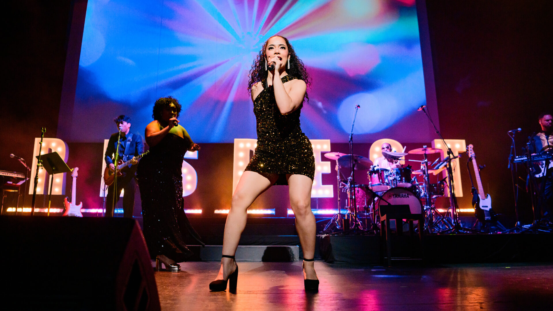 A high-energy live performance on stage featuring a lead singer in a black sequined mini dress and high heels, passionately singing into a microphone. Behind her, a backup singer in a matching black sequined dress and a guitarist accompany the performance. A drummer, bassist, and keyboardist are also visible, illuminated by colorful stage lighting. Large marquee letters spelling 'RESPECT' glow in the background, adding to the vibrant concert atmosphere.