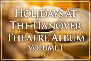 A promotional graphic for "Holidays at The Hanover Theatre Album, Volume I." The text is in white, serif font with a subtle drop shadow, set against a blurred background of warm golden holiday decorations. A thin white border frames the design.