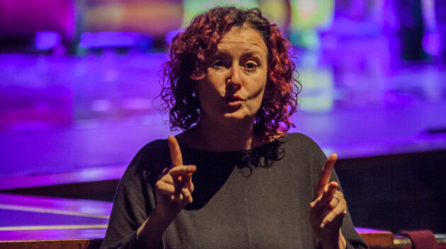 A still image of an ASL interpreter signing during a performance. The interpreter has shoulder length curly brown hair and is wearing a dark turtleneck sweater.