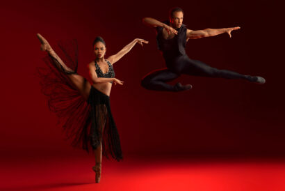 DToH_1920 x 1080 hero image - video card - FB event - Hanover Theatre and Conservatory Two Dance Theatre of Harlem ballet dancers perform against a dramatic red background. The female dancer, in a black fringed outfit, balances en pointe with one leg extended high. The male dancer, dressed in a black sleeveless outfit, leaps mid-air with his arms extended outward.