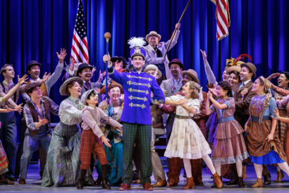 The cast of the Broadway tour of The Music Man celebrates center stage, surrounding Harold Hill in a bright blue band uniform holding a baton. The joyful ensemble is looking to Harold with their hands out toward him, dressed in period costumes.