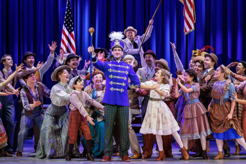 The cast of the Broadway tour of The Music Man celebrates center stage, surrounding Harold Hill in a bright blue band uniform holding a baton. The joyful ensemble is looking to Harold with their hands out toward him, dressed in period costumes.