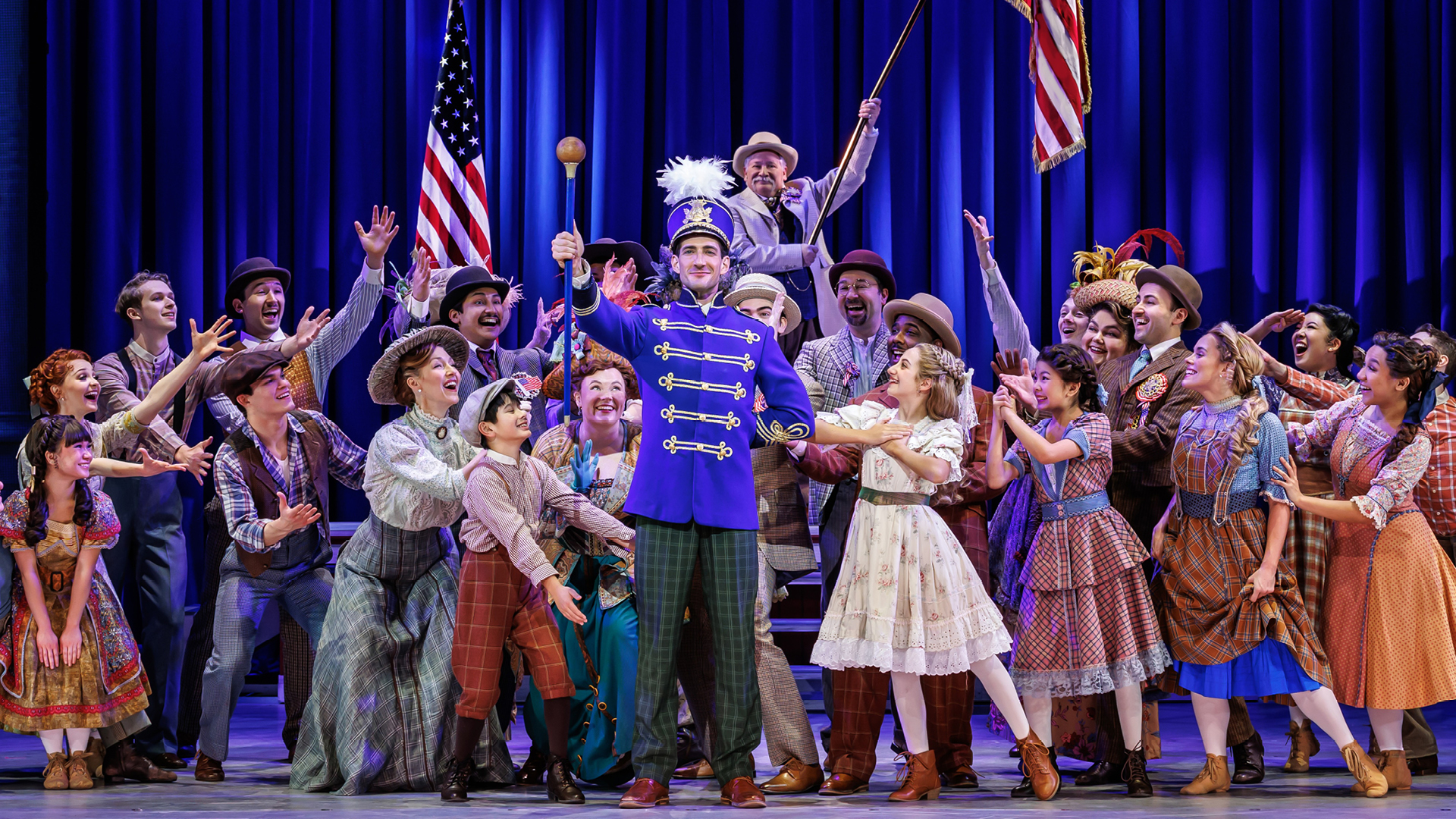 The cast of the Broadway tour of The Music Man celebrates center stage, surrounding Harold Hill in a bright blue band uniform holding a baton. The joyful ensemble is looking to Harold with their hands out toward him, dressed in period costumes.