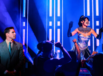 A performer in a sparkling silver corset, fringed skirt and long black gloves sings onstage under vibrant blue and purple lighting as an audience below raises their arms and dance in front of her. To the left, a man in a gray suit and teal tie watches from the side of the stage.