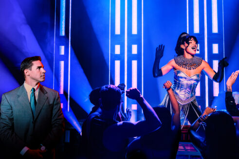 A performer in a sparkling silver corset, fringed skirt and long black gloves sings onstage under vibrant blue and purple lighting as an audience below raises their arms and dance in front of her. To the left, a man in a gray suit and teal tie watches from the side of the stage.