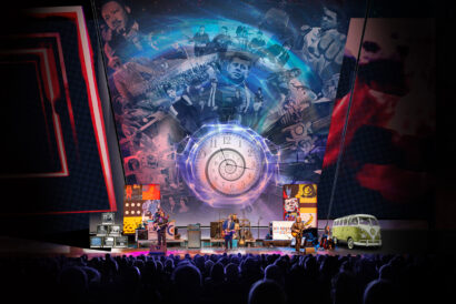 A band performs onstage before a packed audience beneath a large multimedia backdrop featuring a glowing spiral clock and images from the 1960s, including musicians, cultural figures, and a classic Volkswagen bus.