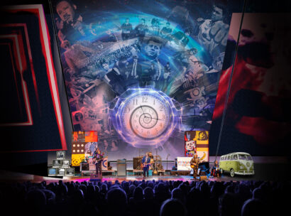 A band performs onstage before a packed audience beneath a large multimedia backdrop featuring a glowing spiral clock and images from the 1960s, including musicians, cultural figures, and a classic Volkswagen bus.