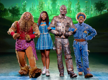 Four cast members from the national tour of The Wiz pose arm-in-arm onstage in colorful costumes, with a glowing Emerald City backdrop behind them.