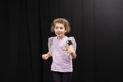 Acting for Commercials Grades 4-7 - Hanover Theatre and Conservatory Young girl with curly hair dances on a dark stage, wearing a lavender top and ruffled sleeves, arms extended.